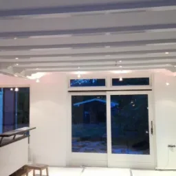 Wnętrze pokoju po malowaniu, z białymi ścianami, dekoracyjnymi belkami sufitowymi i małymi lampami punktowymi, widoczne okna z widokiem na zewnątrz.