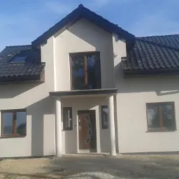 Nowoczesny dom jednorodzinny z ciemną dachówką, widok z przodu na jasnej działce z fragmentem białego busa po lewej stronie.