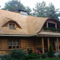 Drewniany dom z unikalnym dachem krytym gontem, z owalnym oknem i balkonem z kwiatami. Widoczne detale wykończenia i naturalne otoczenie.