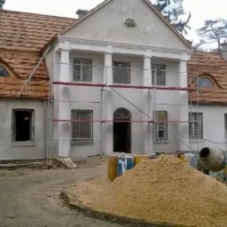 Biały, odrestaurowany dworek z nowym, drewnianym dachem krytym gontem. Widoczne rusztowania, piasek, betoniarka. Prace w toku.