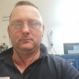 Portret mężczyzny w okularach i ciemnej koszuli polo na tle stanowiska pracy z komputerem i urządzeniami pomiarowymi.