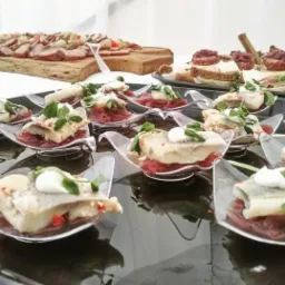 Elegancko podane przekąski: śledź na czerwonej cebuli z kleksem śmietany i listkami ziół, serwowane w przezroczystych, designerskich łyżeczkach na czarnej tacy, w tle deski z wędlinami i kanapkami.