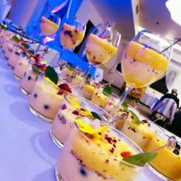 Rząd deserów w szklanych naczyniach i kieliszkach, z dwuwarstwową kompozycją: jasny krem na dole i żółta pianka na górze, udekorowane owocami, płatkami kwiatów i listkami mięty, ustawione na białym...