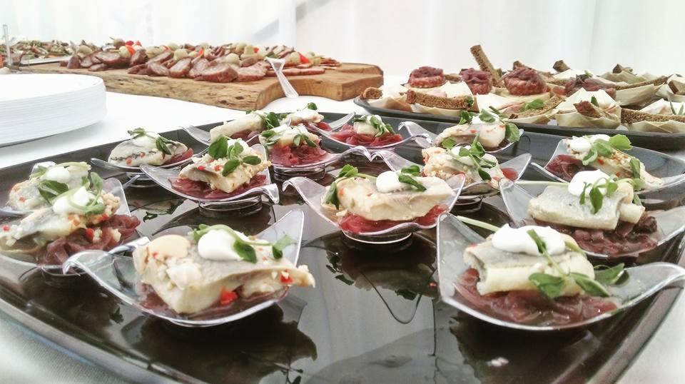 Elegancko podane przekąski: śledź na czerwonej cebuli z kleksem śmietany i listkami ziół, serwowane w przezroczystych, designerskich łyżeczkach na czarnej tacy, w tle deski z wędlinami i kanapkami.