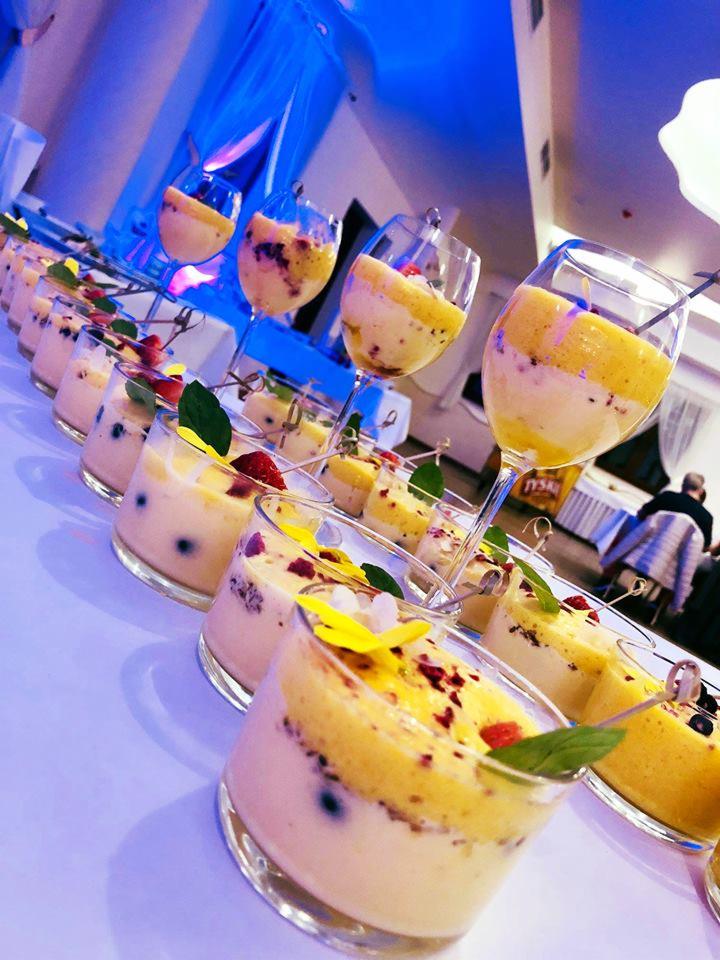 Rząd deserów w szklanych naczyniach i kieliszkach, z dwuwarstwową kompozycją: jasny krem na dole i żółta pianka na górze, udekorowane owocami, płatkami kwiatów i listkami mięty, ustawione na białym...