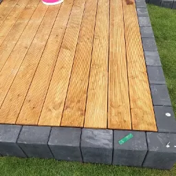 Świeżo zaimpregnowany drewniany taras z ryflowanych desek, obrzeża z szarych betonowych kostek, wiadro z impregnatem Atlas Rapid i listewka na deskach, trawa w tle.