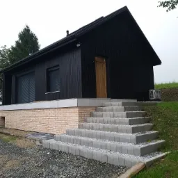 Czarny dom z drewnianą elewacją, jasną cegłą fundamentową i schodami z szarej kostki, widoczny klimatyzator na ścianie