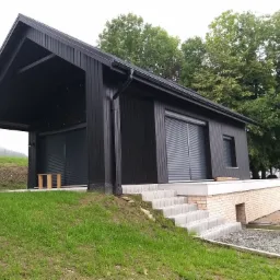 Nowoczesny, czarny dom z drewna z garażem, widoczne schody z kostki brukowej i fragment białego vana z przodu.