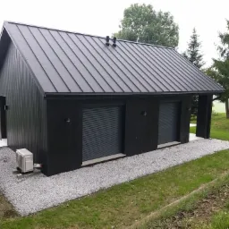 Czarny, parterowy budynek z dwoma garażami z roletami, metalowym dachem i klimatyzatorem na zewnątrz, otoczony trawnikiem i szarym żwirem.