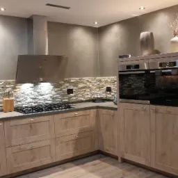 Nowoczesna kuchnia z drewnianymi szafkami, kamienną ścianą nad blatem i stalowym okapem, oświetlona punktowymi lampami sufitowymi i z dekoracyjnymi wazonami na szafce z piekarnikami.