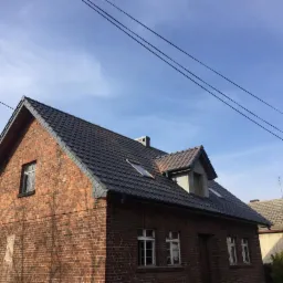 Dom z czerwonej cegły z nowym, ciemnoszarym dachem z dachówki karpiówki i oknami dachowymi, widok na błękitne niebo z przewodami elektrycznymi.
