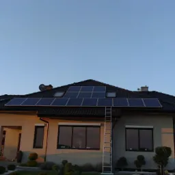 Dom jednorodzinny z panelami słonecznymi na dachu z czarną dachówką. Aluminiowa drabina oparta o dach. Błękitne niebo w tle.