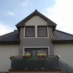 Dwukondygnacyjny dom z balkonem, brązowym dachem i szarą elewacją, z widocznymi oknami z roletami zewnętrznymi i skrzynkami z kwiatami na balustradzie.