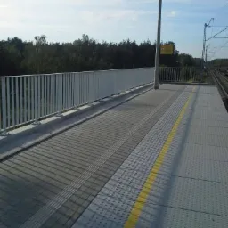 Nowoczesna platforma peronowa z białą metalową balustradą, oświetleniem i żółtą linią bezpieczeństwa, widok na tory kolejowe i zalesiony teren w tle.