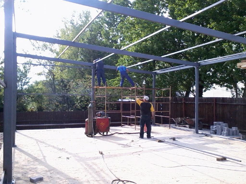 Montaż stalowej konstrukcji szkieletowej z użyciem rusztowań, widoczny spawacz w żółto-czarnej koszulce i butla z gazem spawalniczym na placu budowy.