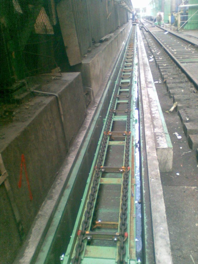 Wnętrze hali produkcyjnej z widocznym systemem transportu łańcuchowego w zagłębieniu posadzki, z elementami konstrukcji stalowej po bokach. Widoczne ślady użytkowania i zabrudzenia.