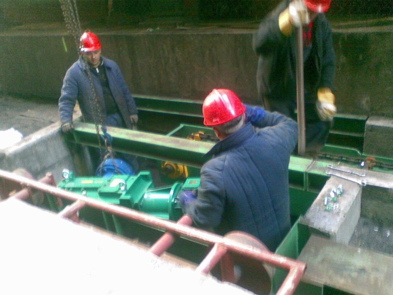 Montaż zielonego agregatu pompowego w stalowej konstrukcji przez pracowników w czerwonych kaskach, widoczne narzędzia i łańcuchy.