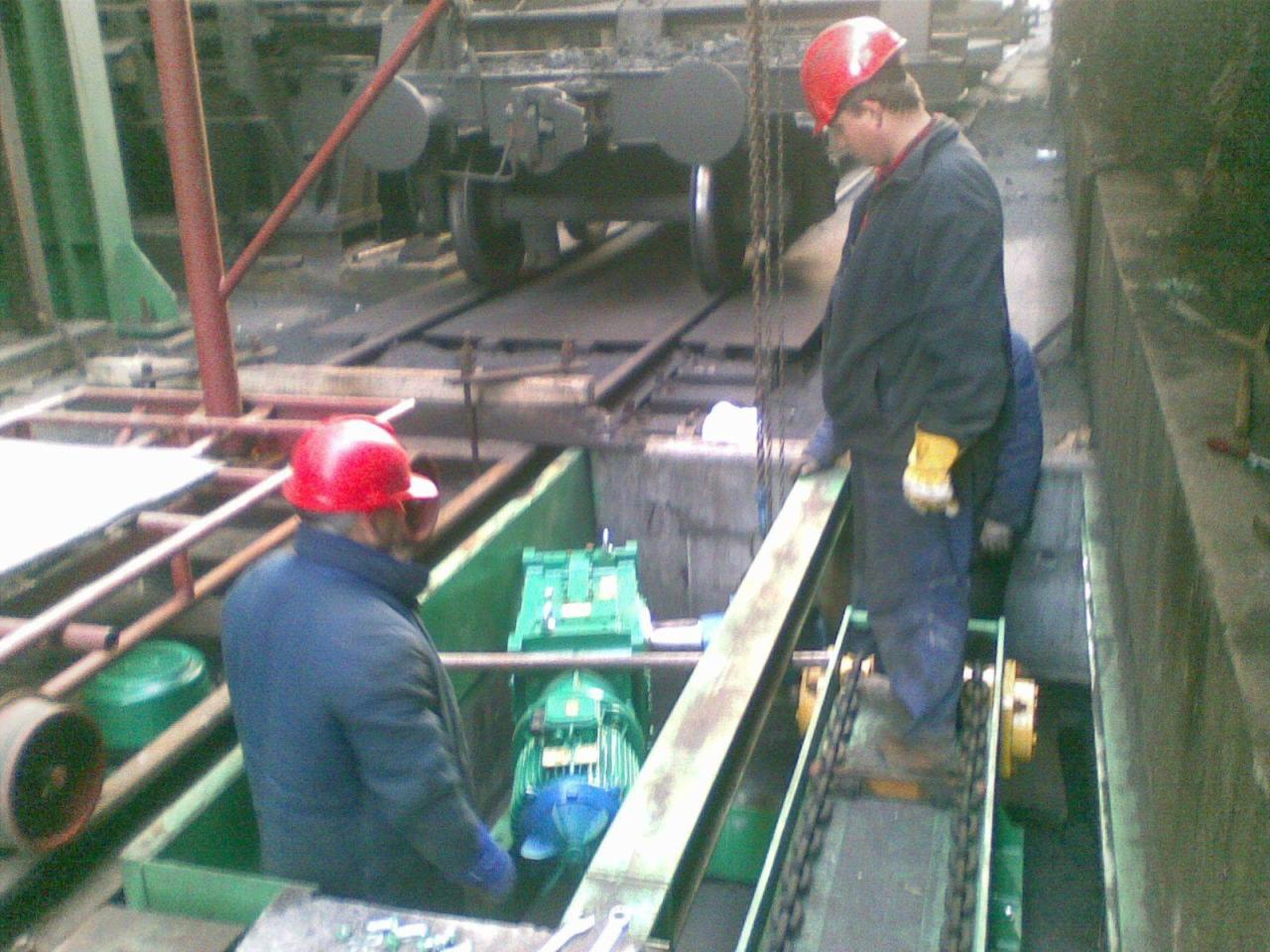 Dwóch pracowników w kaskach i roboczych ubraniach nad zieloną konstrukcją z łańcuchem w hali, w tle wagon kolejowy.