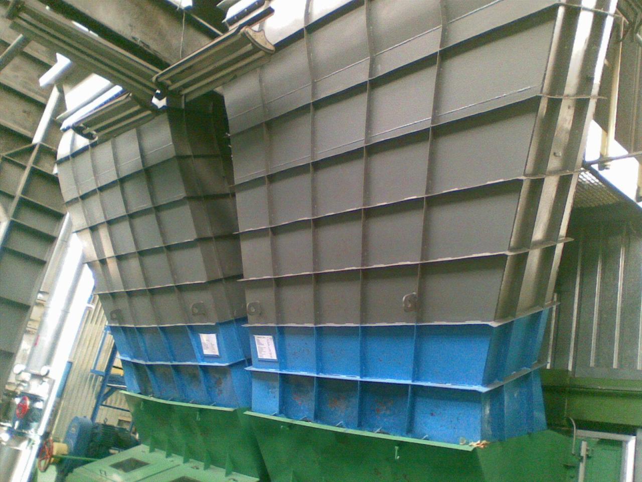 Fragment dużej, pomalowanej na szaro-niebiesko konstrukcji stalowej w hali przemysłowej, z widocznymi spawami i elementami mocującymi.