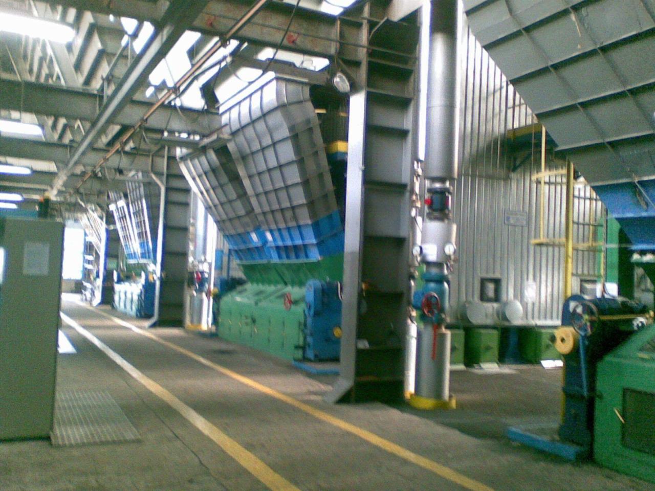 Wnętrze hali produkcyjnej z konstrukcją stalową, widoczne elementy maszyn w odcieniach zieleni i niebieskiego oraz stalowe ramy podtrzymujące dach.