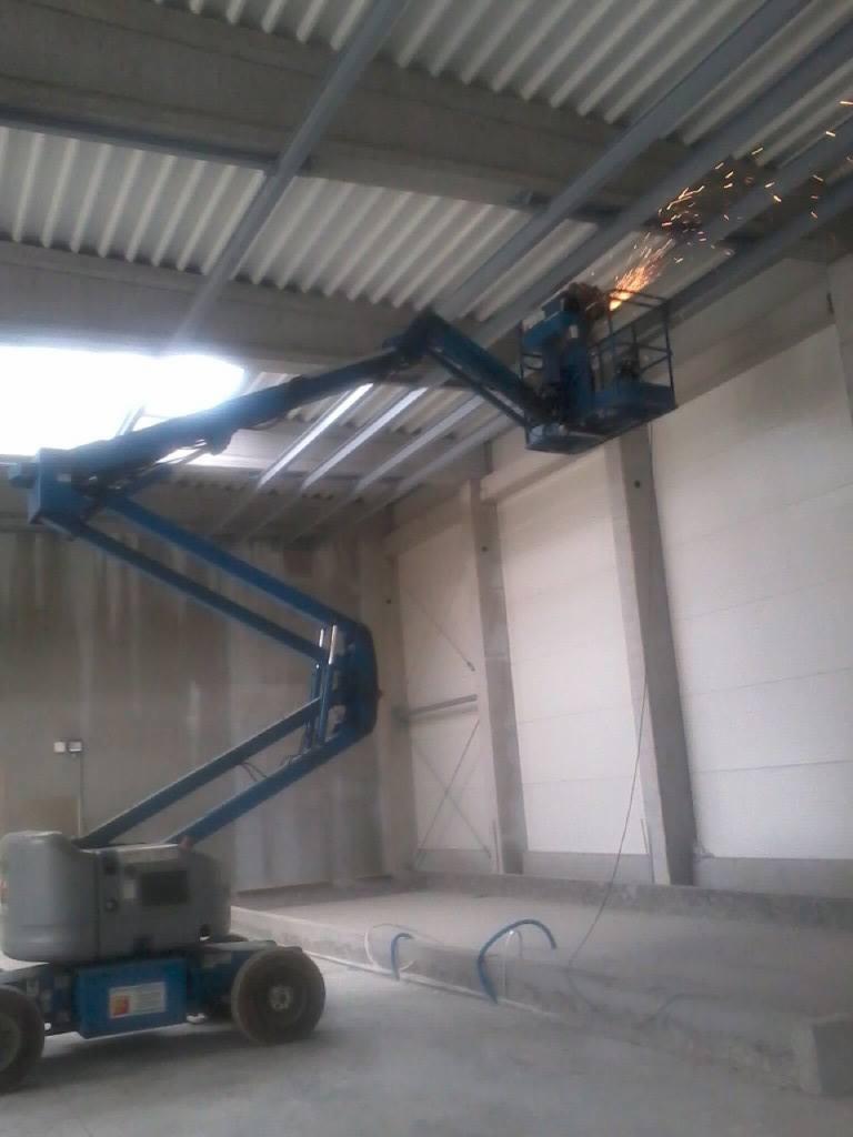 Spawanie konstrukcji stalowej pod dachem hali z podnośnika koszowego, widoczne iskry, surowy stan wykończenia.