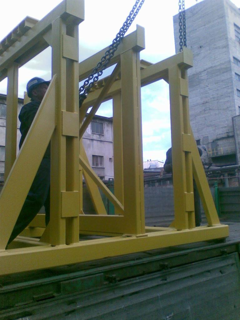 Żółta, modułowa konstrukcja stalowa transportowana na platformie ciężarówki, zabezpieczona łańcuchami, z widocznym pracownikiem w kasku i budynkiem w tle.