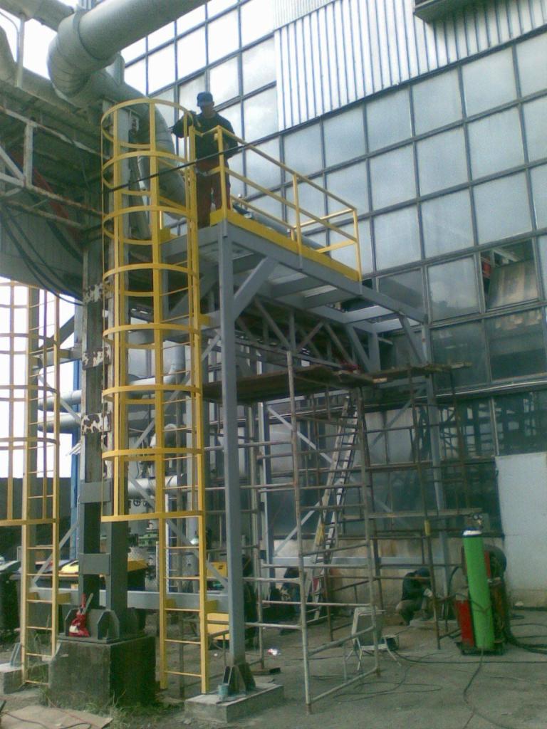 Pracownik na żółtej platformie z drabinką, część konstrukcji stalowej hali przemysłowej z widocznymi oknami i instalacjami.