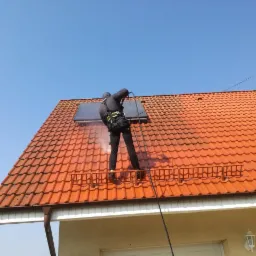 Pracownik w uprzęży, z dyszą ciśnieniową, czyści dach z pomarańczowej dachówki, obok panel słoneczny, widoczna para wodna, na tle błękitnego nieba.