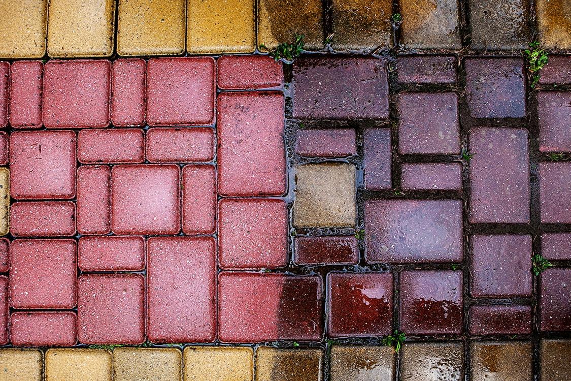 Częściowo umyta kostka brukowa: po lewej stronie czyste, czerwone i żółte kostki, po prawej brudne, mokre, ciemniejsze kostki z drobnymi chwastami w spoinach.