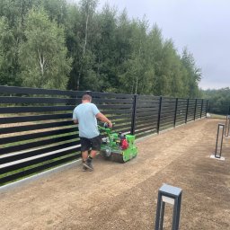FREE-ENERGY SPÓŁKA Z OGRANICZONĄ ODPOWIEDZIALNOŚCIĄ SPÓŁKA KOMANDYTOWA - Pracownik używa aeratora na trawniku wzdłuż nowoczesnego, poziomego ogrodzenia z czarnych paneli. Widoczne lampy ogrodowe i fragment domu z szarą dachówką.