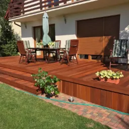 Czerwono-brązowy taras z drewna egzotycznego, z niskimi schodami, wyposażony w stół, krzesła z zielonymi poduszkami i parasol ogrodowy, otoczony trawnikiem i kostką brukową.