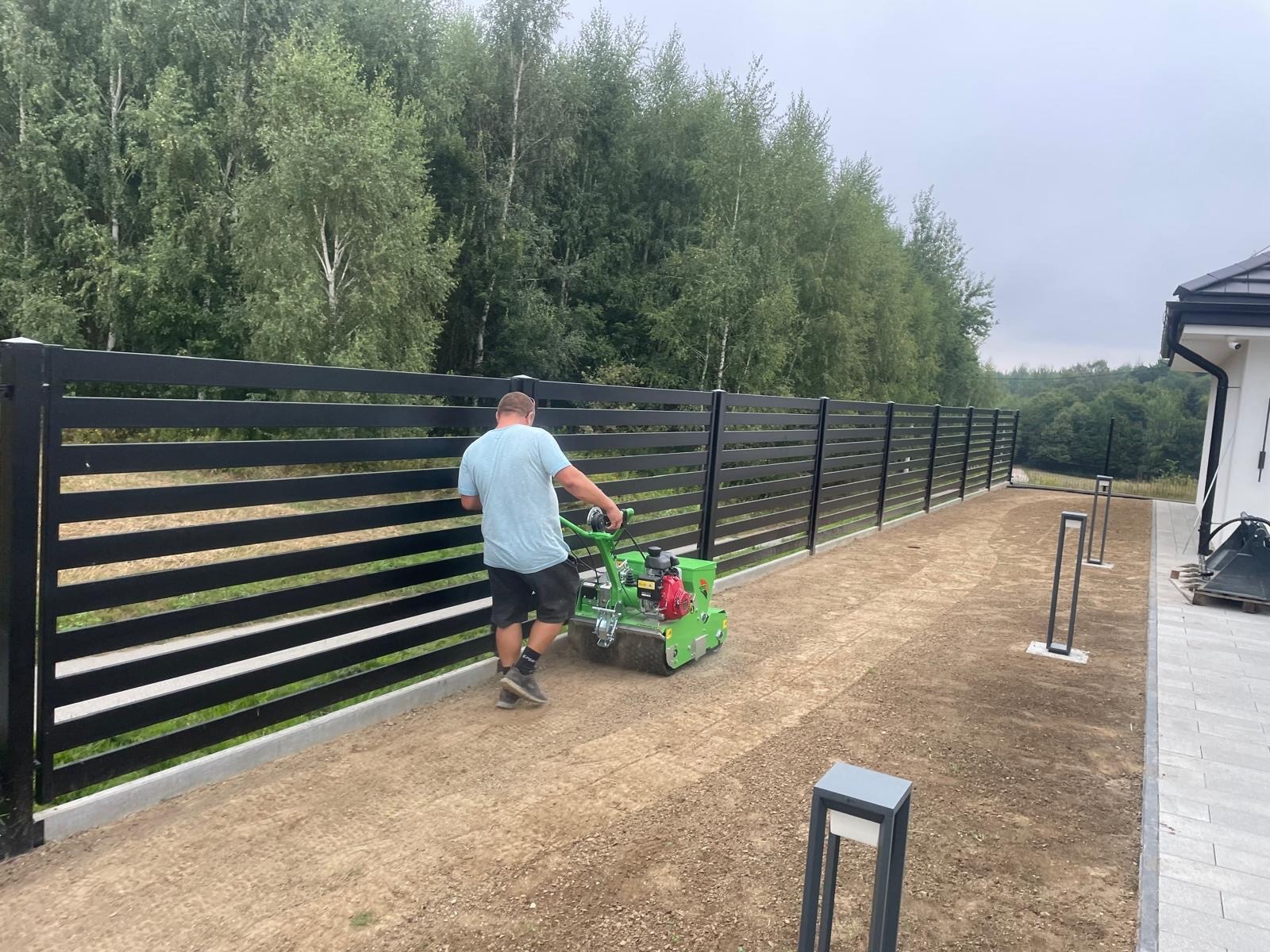 Pracownik używa aeratora na trawniku wzdłuż nowoczesnego, poziomego ogrodzenia z czarnych paneli. Widoczne lampy ogrodowe i fragment domu z szarą dachówką.