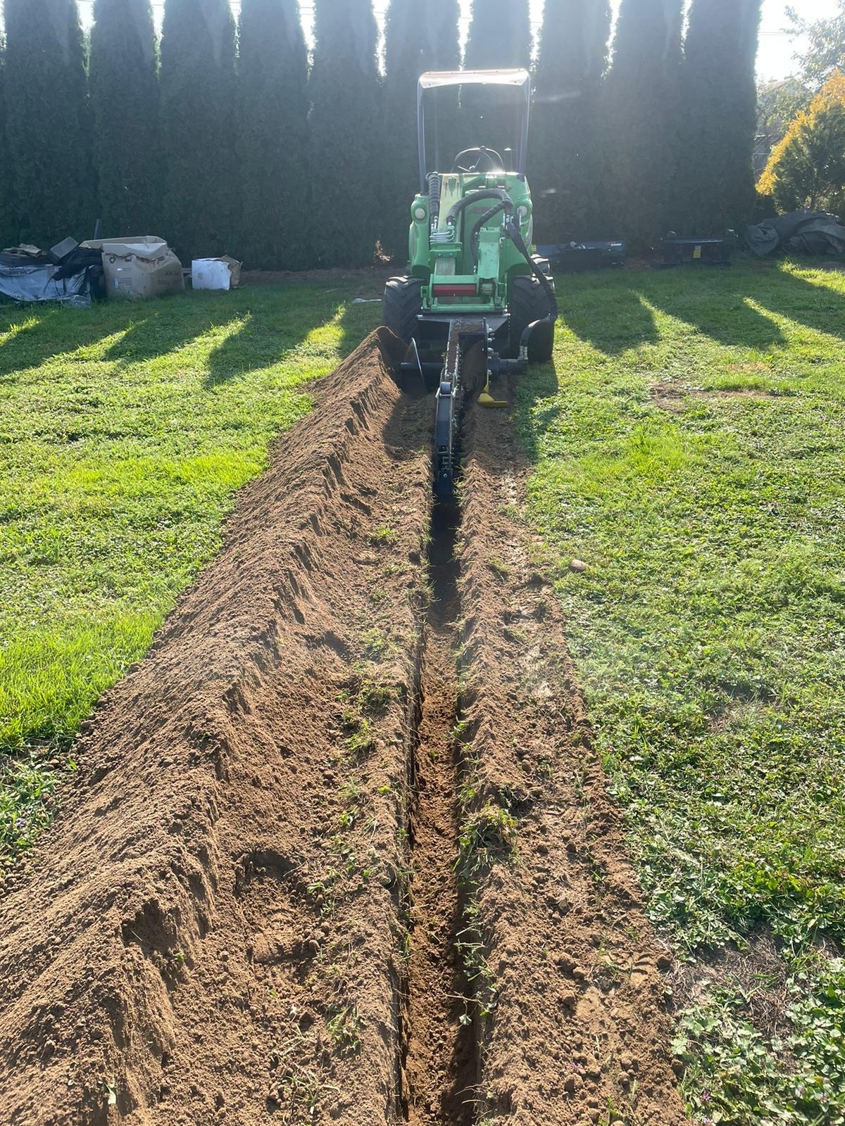 Wąski wykop w trawie wykonany specjalistyczną mini-koparką łańcuchową, widok z góry. Świeżo wykopana ziemia po obu stronach wykopu. Tuje w tle.