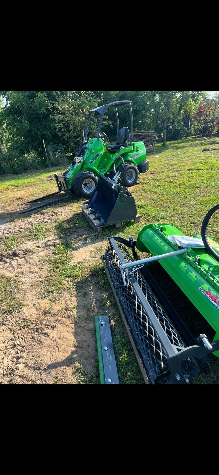 Zielony miniładowarka Avant z łyżką i widłami na palecie, oraz wał do trawy na trawiastym terenie. Sprzęt gotowy do pracy.