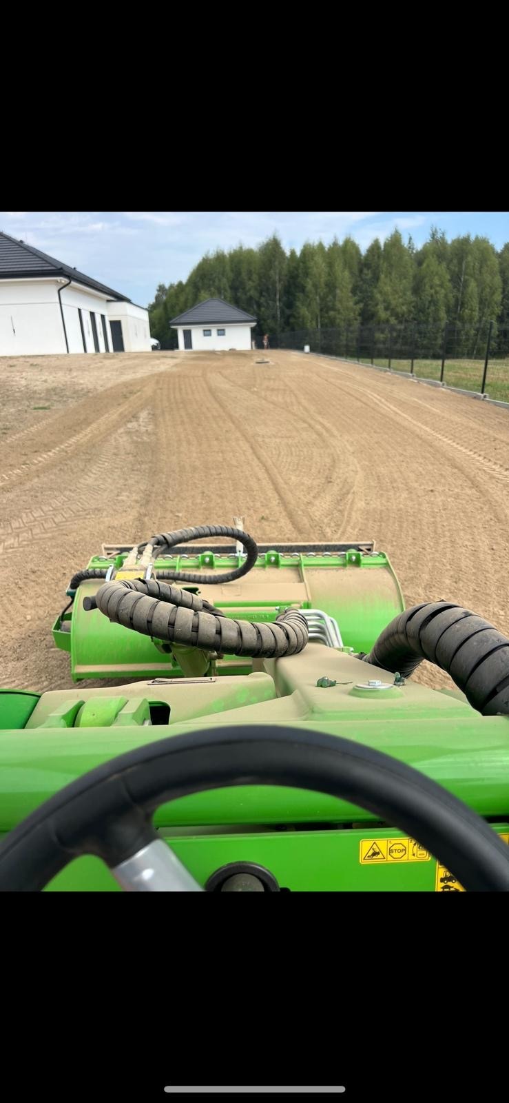Widok z perspektywy operatora na wyrównany teren pod budowę, z widocznymi śladami maszyny, ogrodzeniem i budynkami w tle. Maszyna w kolorze zielonym.