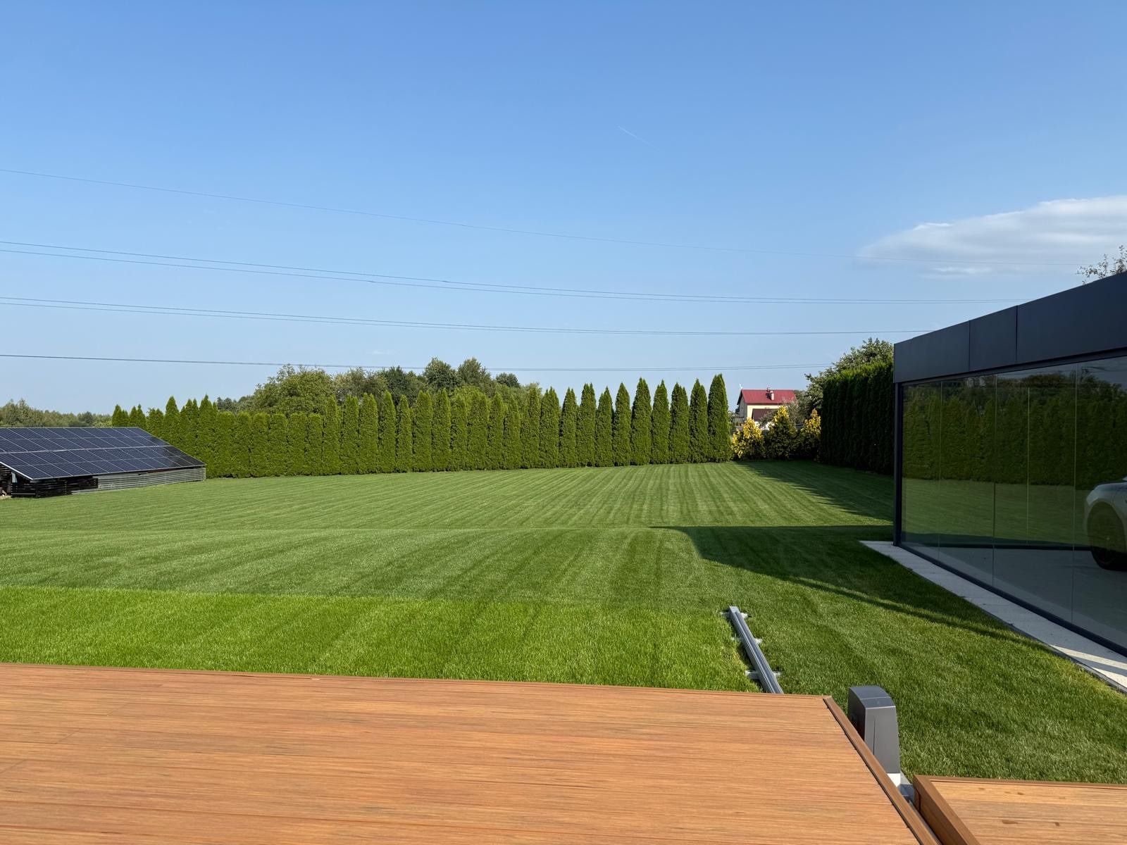 Perfekcyjnie skoszony trawnik z równymi pasami, otoczony tujami i nowoczesnym budynkiem z przeszkloną ścianą. W tle panele słoneczne i dom z czerwonym dachem.