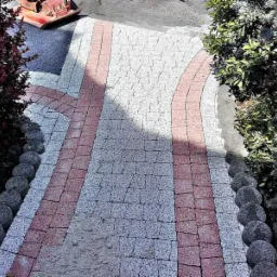 Ścieżka z kostki brukowej w odcieniach szarości i r&oacute;żu, obrzeża wykończone okrągłymi kamieniami, z boku widoczne rośliny i ubijarka w tle.