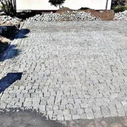 Ułożona nawierzchnia z kostki granitowej w odcieniach szarości, z widocznym stosem kostki do dalszej pracy oraz fragmentem trawnika w tle.