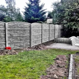 Montaż ogrodzenia z prefabrykowanych paneli betonowych imitujących kamień, widoczne elementy składowane na palecie, trawnik z fragmentami ziemi po pracach ziemnych.
