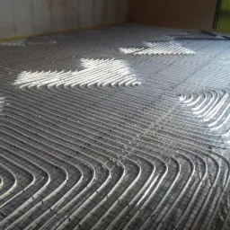 Szare rury ogrzewania podłogowego rozłożone na folii, przymocowane do siatki z drutu, widoczne promienie słoneczne