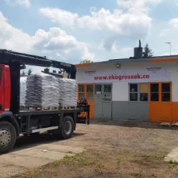 Czerwona ciężarówka z dźwigiem załadowana workami, na tle budynku z banerem reklamowym i stosem palet. Dzień.