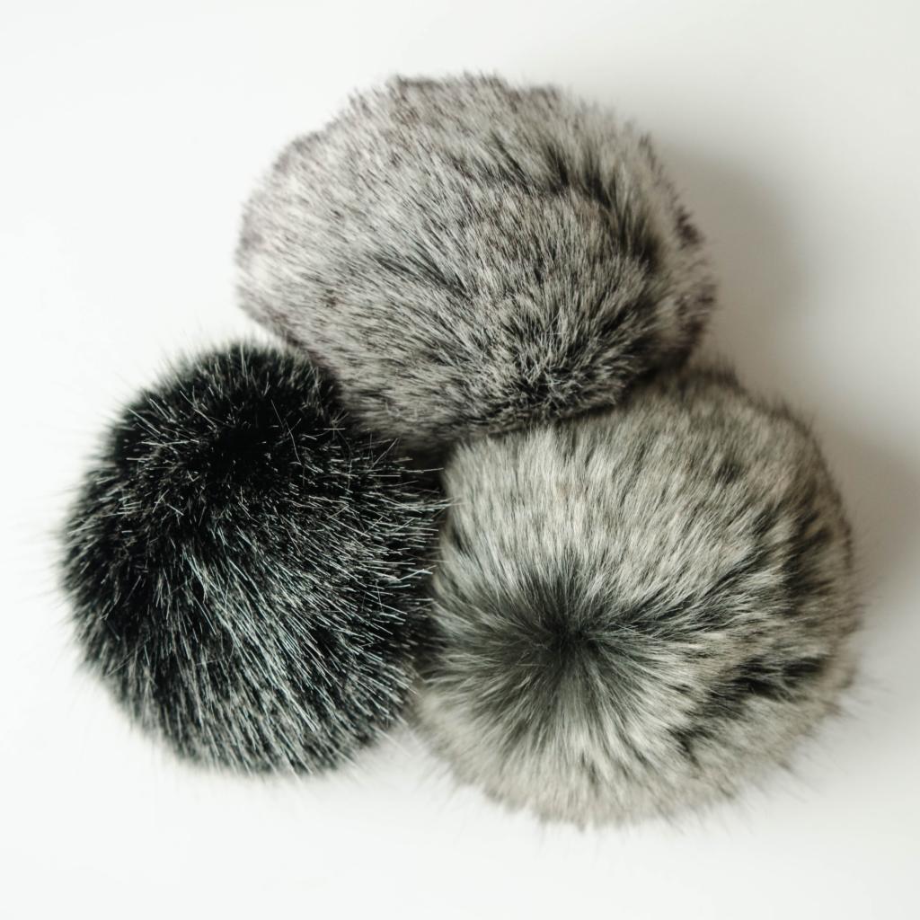 Trzy futrzane pompony w odcieniach szarości i czerni, ułożone w trójkąt na białym tle, prezentujące różne rodzaje wykończeń futrzanych.
