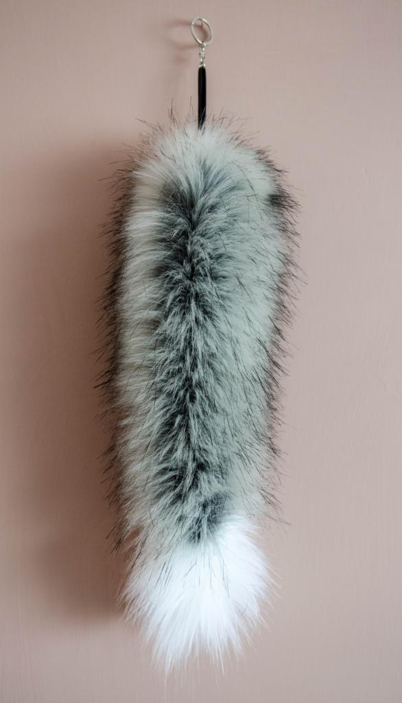Puszysty brelok z imitacji futra w odcieniach szarości i bieli, zawieszony na srebrnym kółku z czarnym paskiem, na tle jednolitej, różowej ściany.