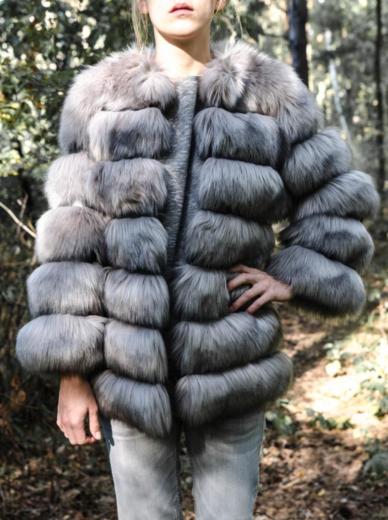 Modelka w szarym futrze z lisów na tle leśnej scenerii. Unikalny design futra z poziomymi pasami. Stonowana kolorystyka i naturalne światło.