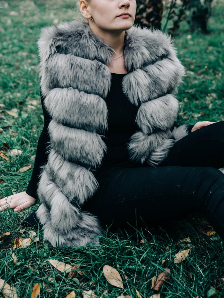 Kobieta w czarnym ubraniu i futrzanym szalu w odcieniach szarości siedzi na trawie z opadłymi liśćmi, spoglądając w górę.