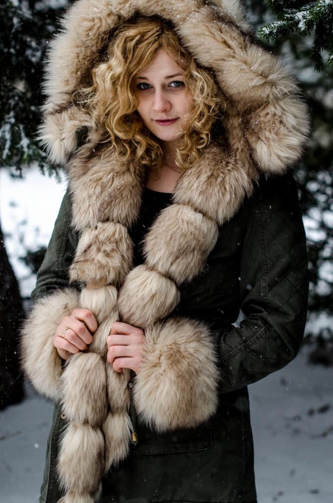 Kobieta w zimowej kurtce z futrzanym kapturem i wykończeniami, trzymająca futrzany szal. Zimowa sceneria z opadającym śniegiem w tle.