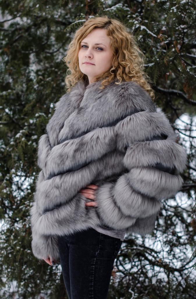 Kobieta w futrzanej, szarej kurtce na tle zimowego krajobrazu. Kręcone blond włosy, dżinsy. Ujęcie od pasa w górę, lekko z dołu. Widoczna struktura futra kurtki.