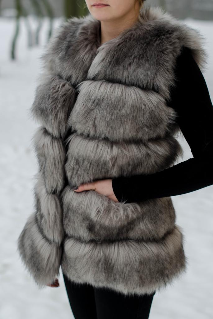 Kobieta w futrzanej kamizelce w odcieniach szarości, stojąca na tle zimowego krajobrazu. Kamizelka ma poziome pasy futra i sięga do bioder.