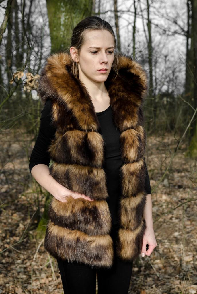 Kobieta w futrzanej kamizelce z kapturem na tle leśnym. Kamizelka w odcieniach brązu i beżu. Ujęcie portretowe, z ręką w kieszeni. Stylizacja jesienna.