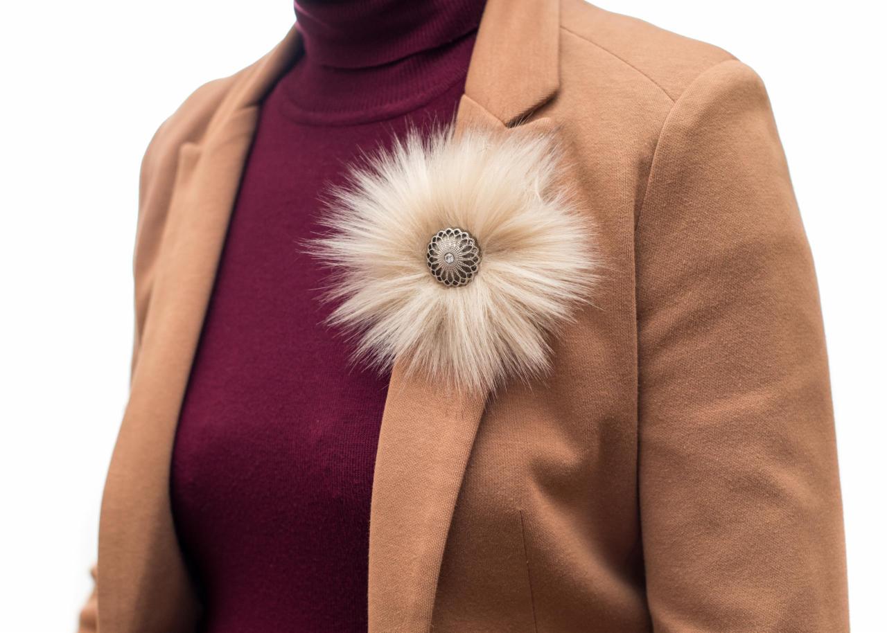 Osoba w karmelowym żakiecie z futrzaną broszką w kształcie kwiatu z ozdobnym, metalowym środkiem, ubrana na bordowy golf.